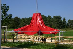 Rotes, zeltartiges Pavillon-Dach, das an einem Kran hängt, über einer Holzstruktur in einer grünen Parklandschaft