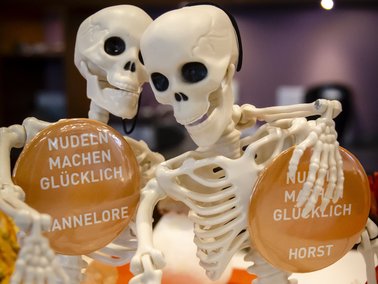 Zwei Skelettfiguren halten jeweils ein rundes Schild mit den Texten 'NUDELN MACHEN GLÜCKLICH' und den Namen 'ANNELORE' sowie 'HORST'.