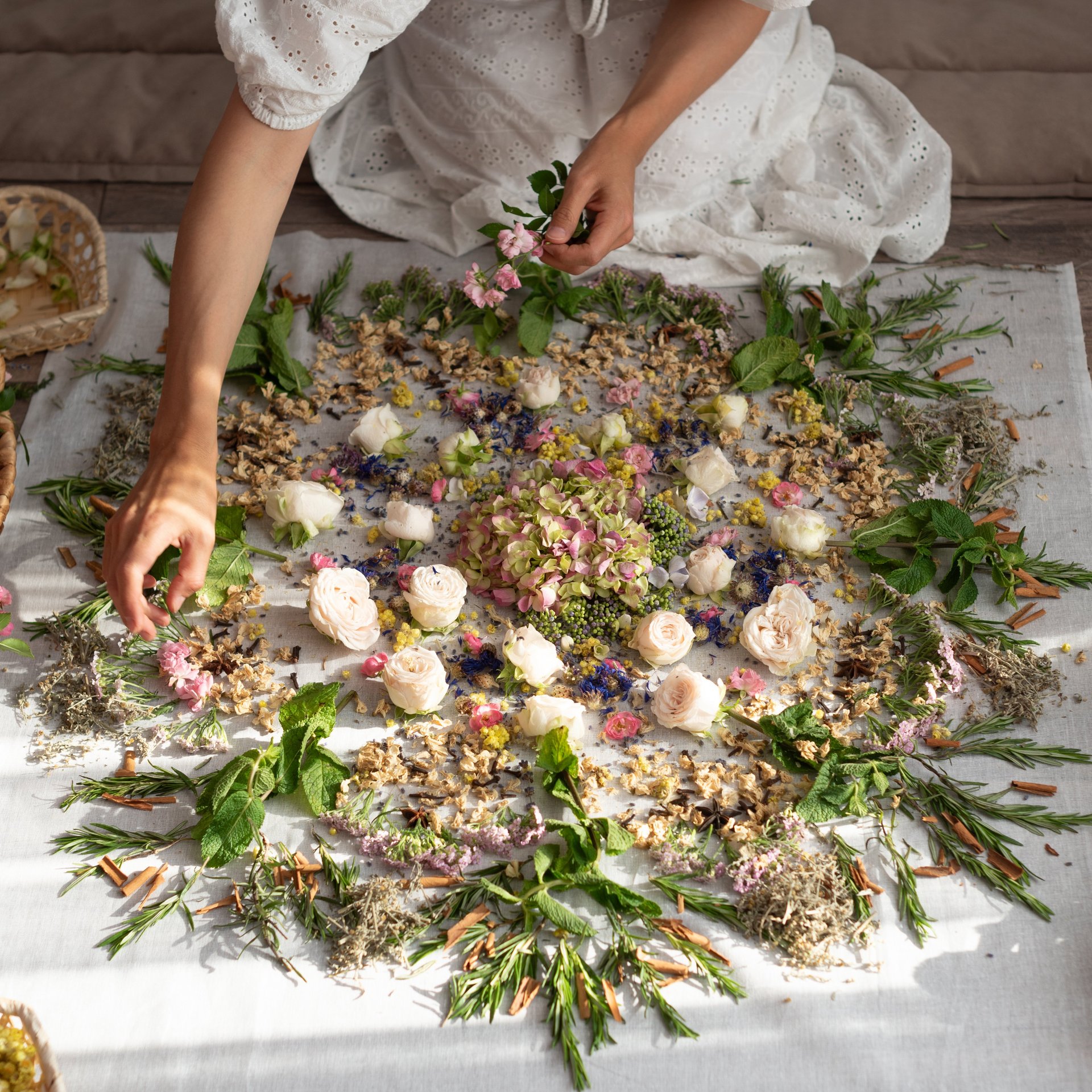 Frau in weißem Kleid arrangiert Blumen und Blätter auf weißem Untergrund in kreisförmigem Muster.