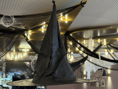 Schwarzer Hexenhut mit breiter Krempe und spitz zulaufender Spitze, hängend in einem Raum mit Deckenbeleuchtung.