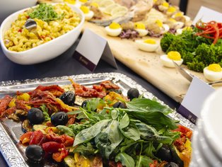 Buffet mit verschiedenen Salaten, darunter grüner Blattsalat, schwarze Oliven, getrocknete Tomaten und Eierhälften auf Holzbrett.