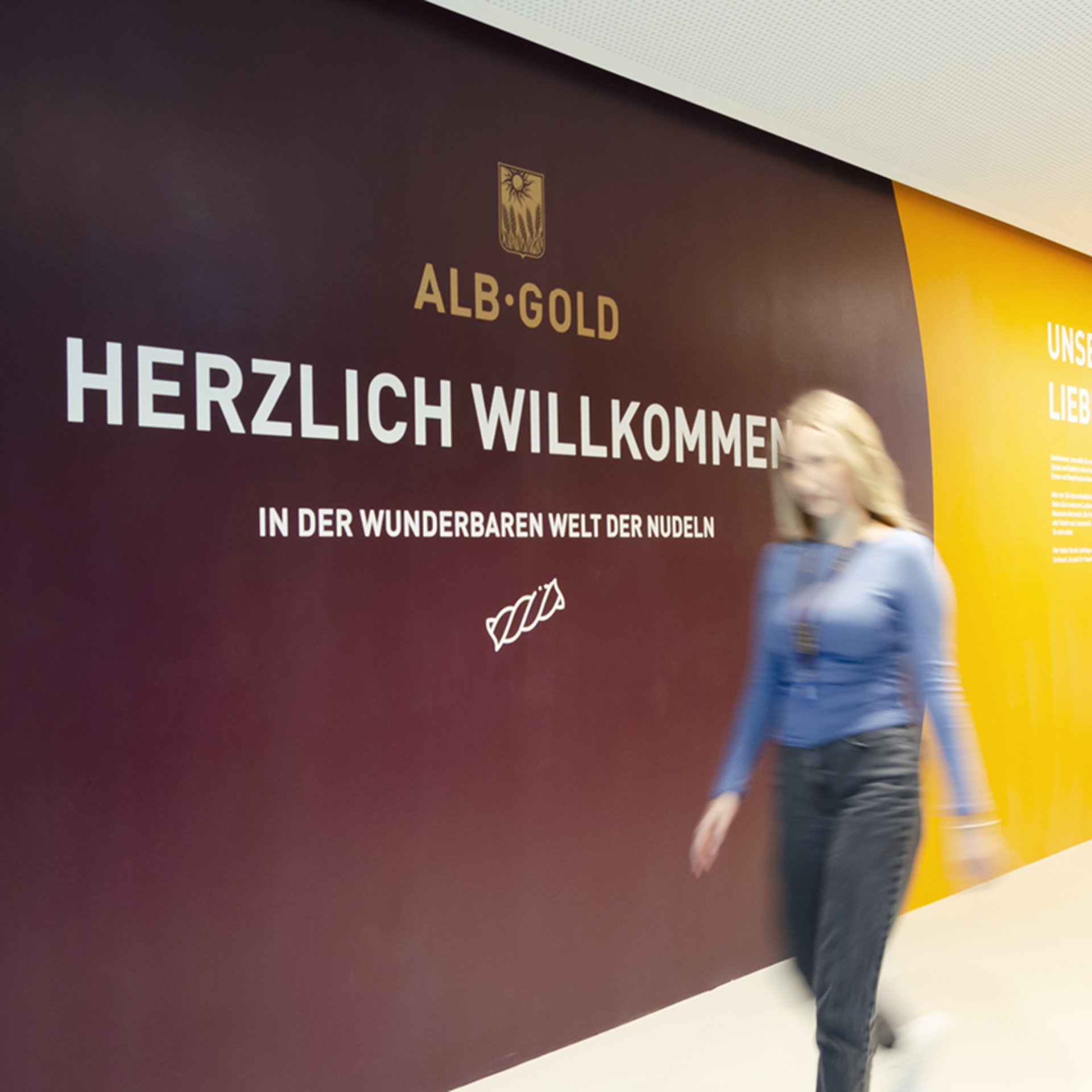 Wand mit Schrift 'ALB-GOLD HERZLICH WILLKOMMEN IN DER WUNDERBAREN WELT DER NUDELN' und Logo über dem Text