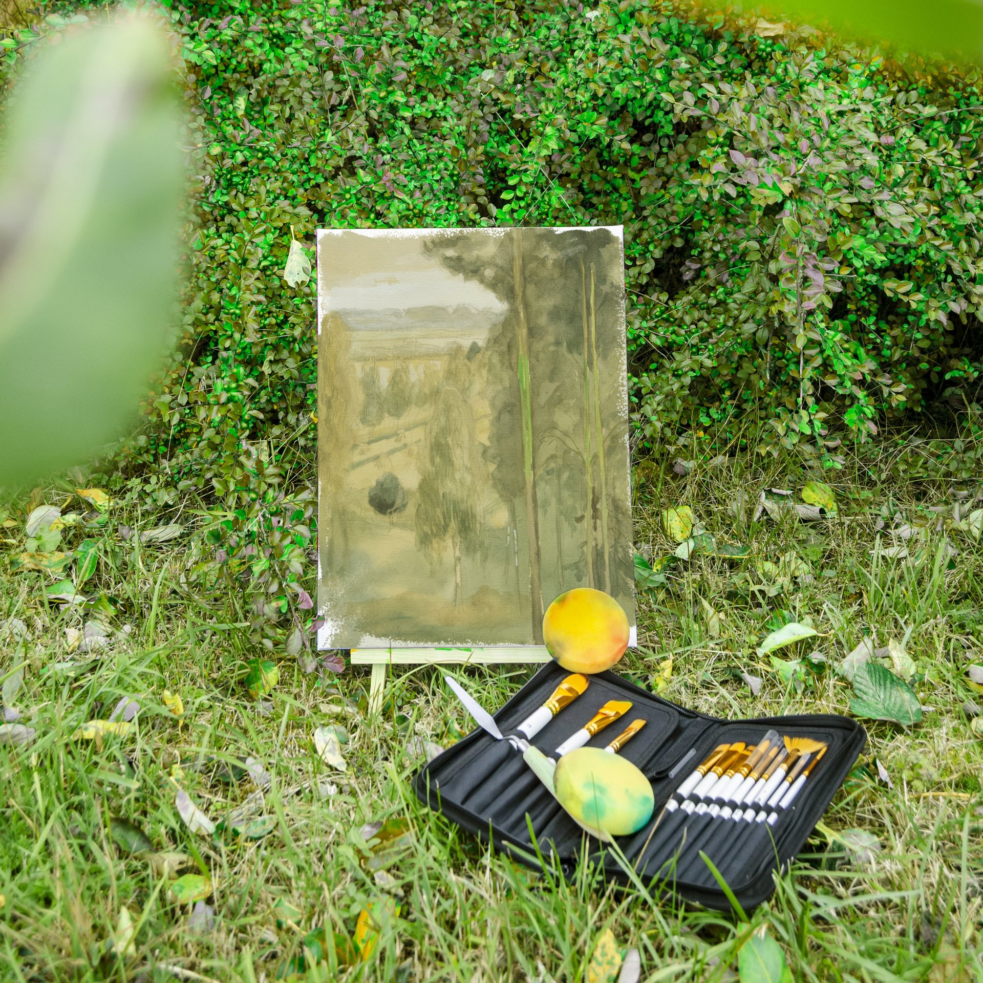 Offenes schwarzes Malkasten mit Pinseln und Farben auf Gras, dahinter Staffelei mit Leinwand vor grünem Busch.
