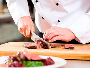 Koch in weißer Jacke schneidet auf Holzbrett ein Stück gebratenes Fleisch