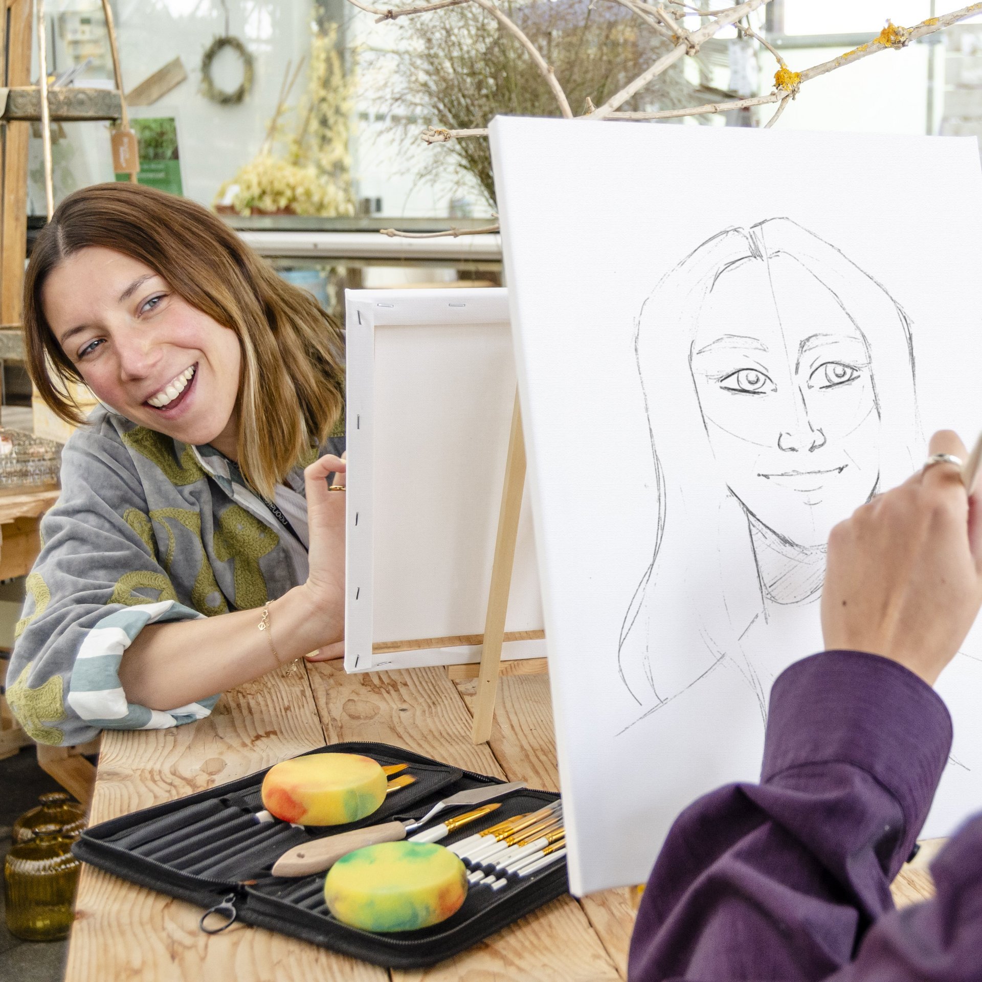 Person zeichnet mit Bleistift auf Skizzenblock, daneben liegen zwei Schwämme und Pinsel auf einem Tisch.