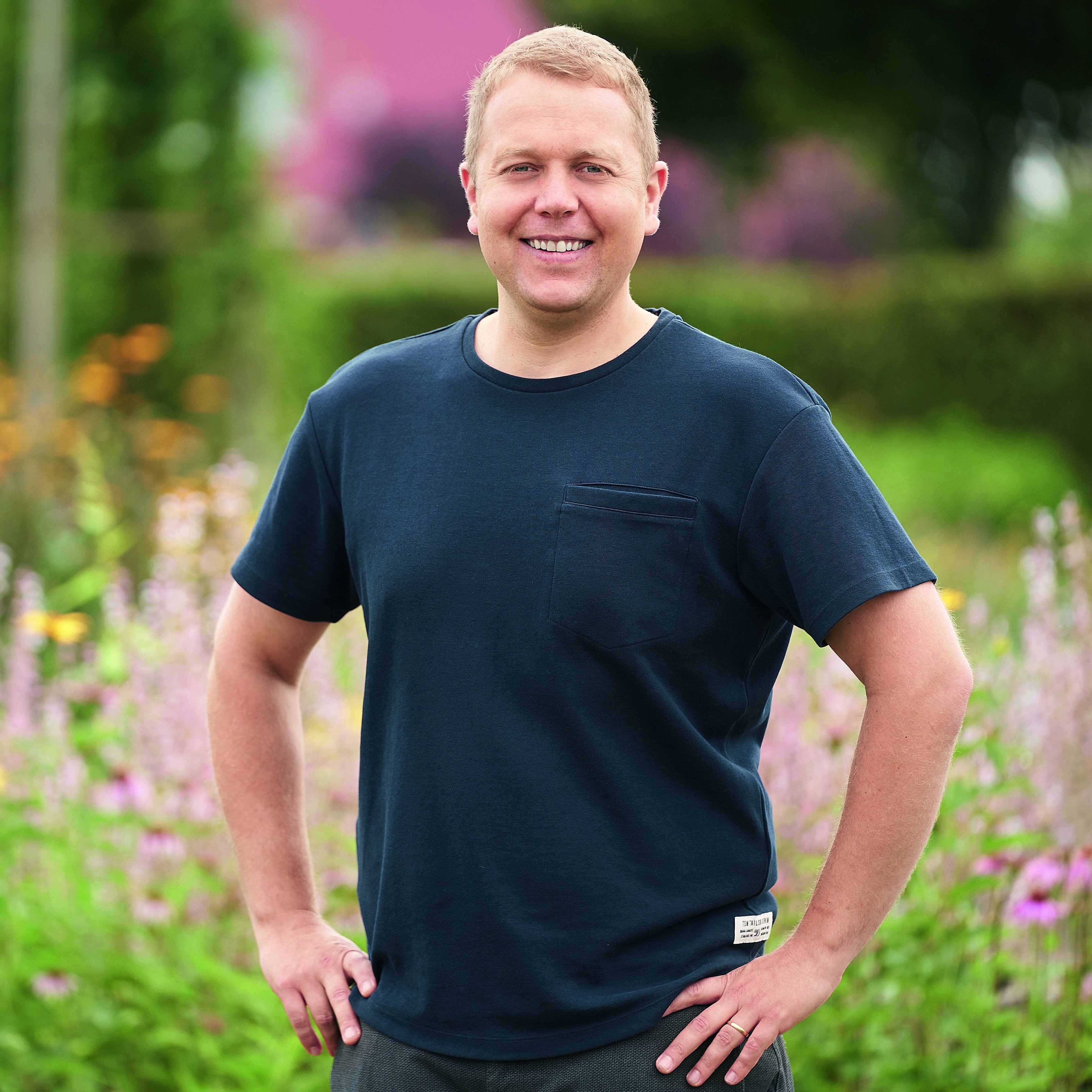 Mann steht mit Händen in der Hüfte in einem Garten mit bunten Blumen und grünen Pflanzen