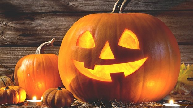 Leuchtender Halloween-Kürbis mit ausgeschnittenen Augen, Nase und lachendem Mund, umgeben von weiteren Kürbissen auf Holzboden vor Holzwand.