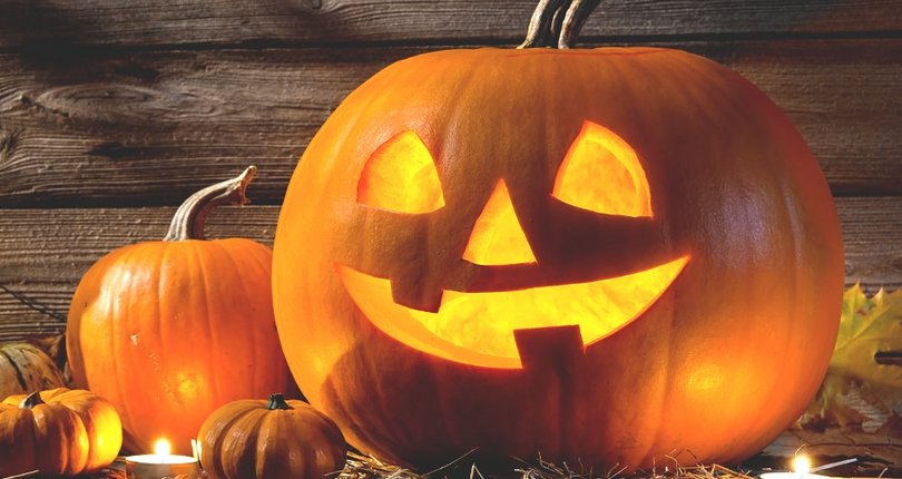 Leuchtender Halloween-Kürbis mit ausgeschnittenen Augen, Nase und lachendem Mund, umgeben von weiteren Kürbissen auf Holzboden vor Holzwand.