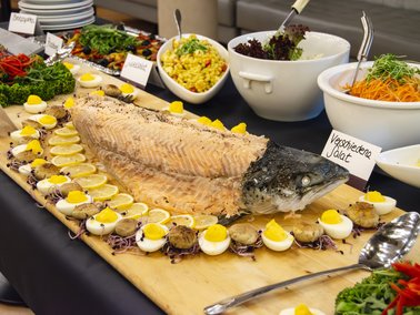Ganz ausgenommenes Fischfilet mit Kopf auf Holzbrett, umgeben von halbierten Eiern mit Eigelb und mehreren Schüsseln mit Salaten im Hintergrund.