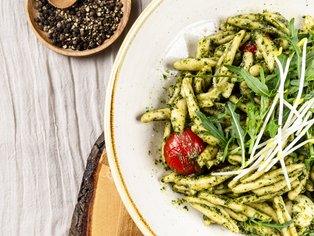 Teller mit grünem Pesto-Pasta, Kirschtomate und Sprossen, daneben Holzunterlage und Schale mit Pfefferkörnern.