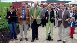 Fünf Personen schneiden gemeinsam ein Band durch bei der Einweihung im Rahmen des Sommermarktes, umgeben von Zuschauern und grüner Landschaft.