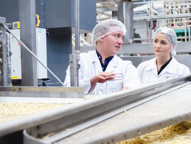 Drei Personen in weißen Schutzkitteln und Haarnetzen stehen an einer Produktionslinie mit Nudeln in einer Nudelproduktionsanlage.