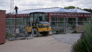 Baustelle des Kräutermarktes mit einem gelben Baumaschinenfahrzeug vor einem modernen Glasgebäude im Bau