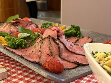 Aufgeschnittenes Roastbeef auf einem Holzbrett mit Tomaten und Brokkoli auf einem rot-weiß karierten Tischtuch