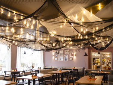 Gastraum eines Restaurants mit mehreren Holztischen, schwarzen Stühlen und einer Decke, die mit schwarzen Netzen und Lichterketten dekoriert ist.