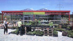 Kräutermarktstand mit Pflanzen auf Paletten vor einem roten Metallgerüst mit grünem Schild 'Kräuter Garten Markt'