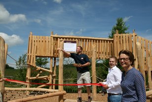 Drei Personen bei der Eröffnung eines Holzspielplatzes, eine hält ein Schild, eine durchschneidet ein rotes Band