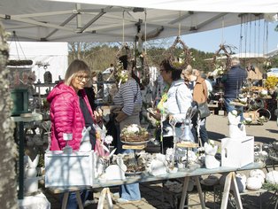 Marktstand mit dekorativen Keramikfiguren, Körben und Blumen unter einem weißen Pavillon, mehrere Personen betrachten die Auslage