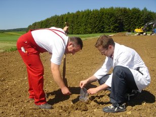 Zwei Männer untersuchen den Boden auf einem Acker, einer trägt rote Arbeitshosen und hält einen Spaten, der andere kniet und hält eine Bodenprobe Zwei Männer untersuchen den Boden auf einem Acker, einer trägt rote Arbeitshosen und hält einen Spaten, der andere kniet und hält eine Bodenprobe