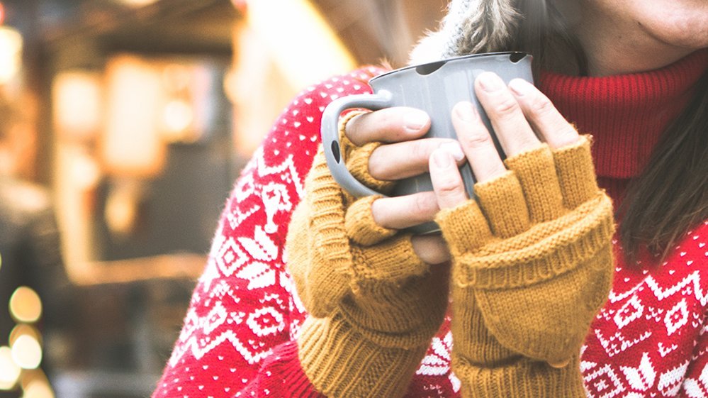 Frau in rotem Norwegerpullover und gelben Handschuhen hält graue Tasse mit beiden Händen