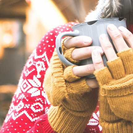 Frau in rotem Norwegerpullover und gelben Handschuhen hält graue Tasse mit beiden Händen