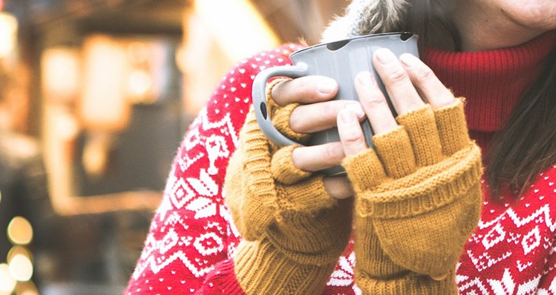 Frau in rotem Norwegerpullover und gelben Handschuhen hält graue Tasse mit beiden Händen