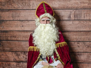 Person in Nikolaus-Kostüm mit rotem Bischofsmantel, weißem Bart und roter Mitra vor Holzwand stehend.