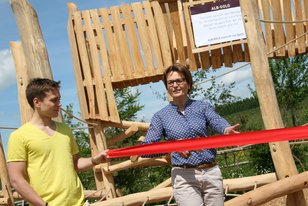 Zwei Personen schneiden bei der Eröffnung eines Spielplatzes ein rotes Band vor einem Holzspielgerät durch