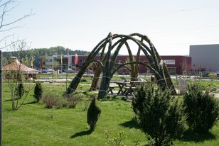 Weidenpavillon aus gebogenen Weidenruten auf einer grünen Wiese mit Bänken darunter, umgeben von kleinen Sträuchern und Gebäuden im Hintergrund