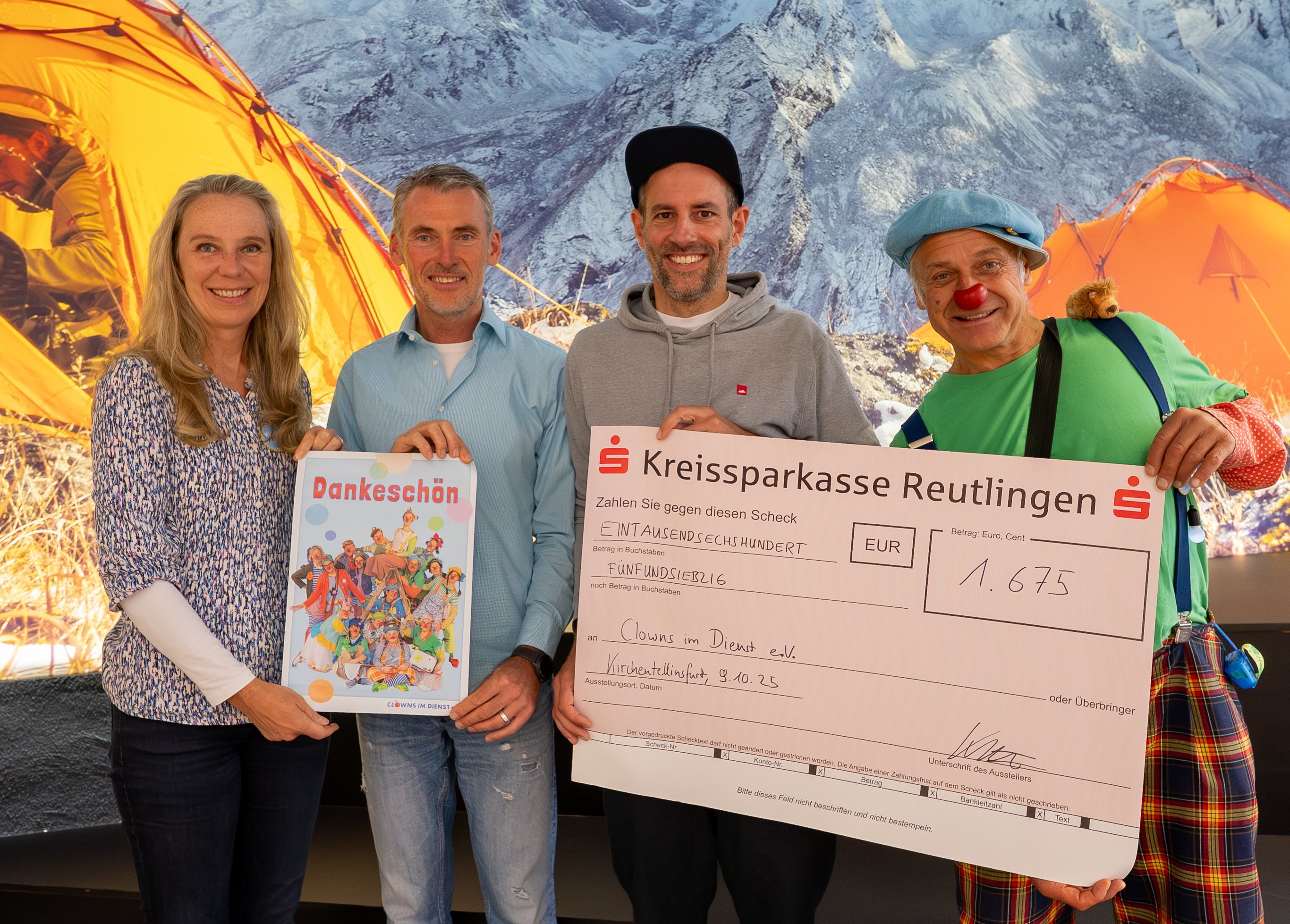 Vier Personen stehen vor Bergkulisse mit zwei orangefarbenen Zelten, eine hält ein buntes Plakat, eine einen großen Scheck der Kreissparkasse Reutlingen.