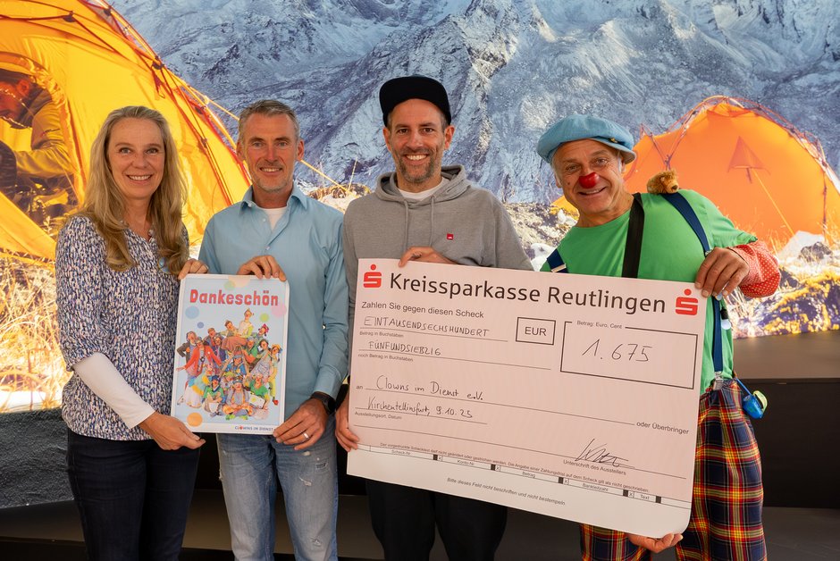 Vier Personen vor Bergkulisse mit orangefarbenen Zelten, zwei halten einen großen Scheck der Kreissparkasse Reutlingen.