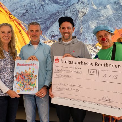 Vier Personen stehen vor Bergkulisse mit zwei orangefarbenen Zelten, eine hält ein buntes Plakat, eine einen großen Scheck der Kreissparkasse Reutlingen.