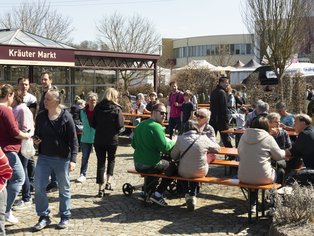 Menschen sitzen und stehen an Tischen vor einem Marktstand mit dem Schild 'Kräuter Markt' unter freiem Himmel an einem sonnigen Tag