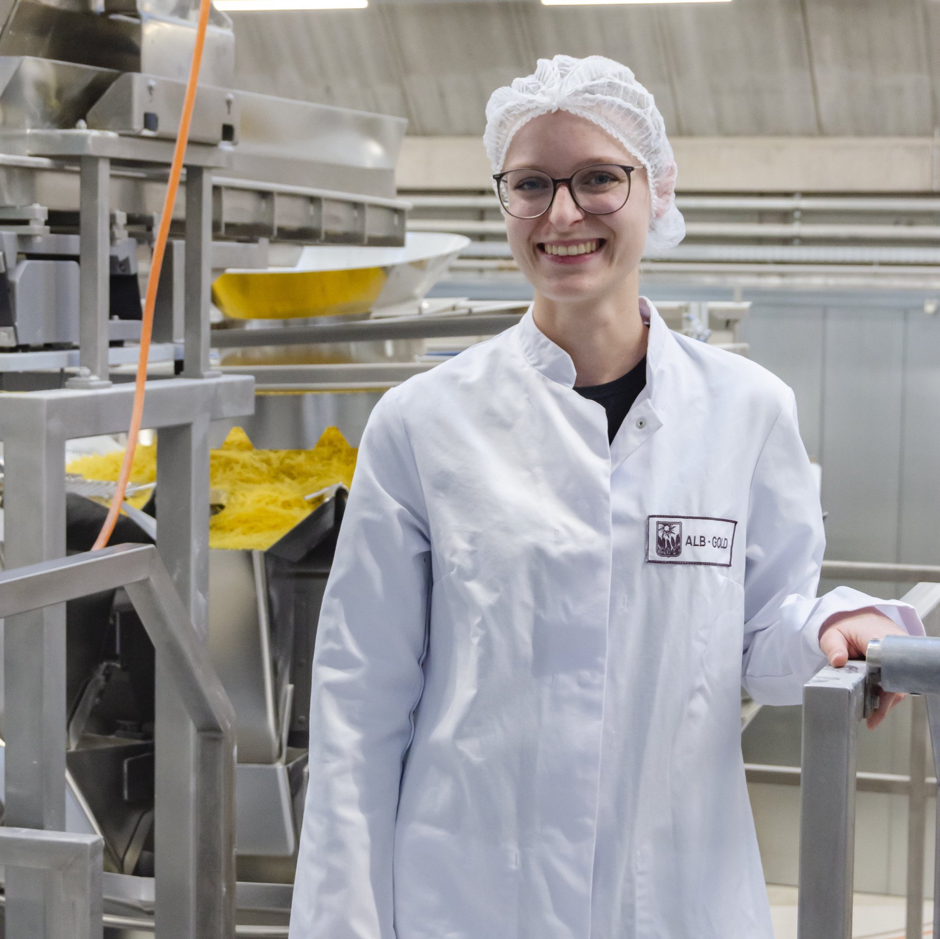 Person in weißem Laborkittel und Haarnetz steht in industrieller Produktionsumgebung und hält sich am Geländer fest.