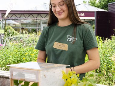 Frau mit grünem T-Shirt steht an einem Tisch im Garten, hält ein kleines Terrarium mit Pflanzen