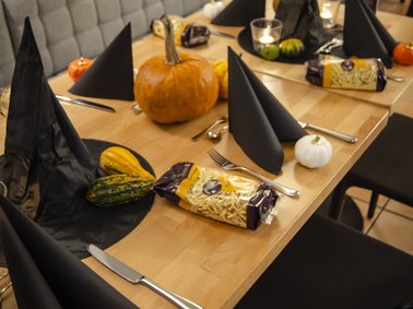 Langer Holztisch mit schwarzem Besteck, schwarzen Papierservietten, mehreren Kürbissen und verpacktem Brot, umgeben von schwarzen Stühlen und grauem Polstersofa.