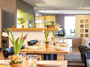 Restaurant mit gedeckten Holztischen, schwarzen Stühlen und Blumen in Vasen auf den Tischen.