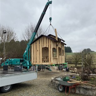 Kran hebt ein hölzernes Spielhaus auf einem Spielplatz während der Eröffnung, ländliche Umgebung mit Bäumen und Wiese im Hintergrund
