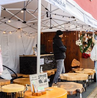 Marktstand mit Holzscheiben als Tische und Dekoration, davor stehen mehrere Personen in Winterkleidung auf einem Bürgersteig