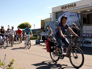 Eine Gruppe von Fahrradfahrern fährt an einem ALB-GOLD Verkaufsstand auf einer Straße vorbei.