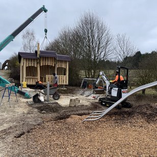 Kran hebt ein hölzernes Spielhaus auf einem Spielplatz mit einer Rutsche während der Eröffnung, ländliche Umgebung mit Bäumen und Wiese im Hintergrund
