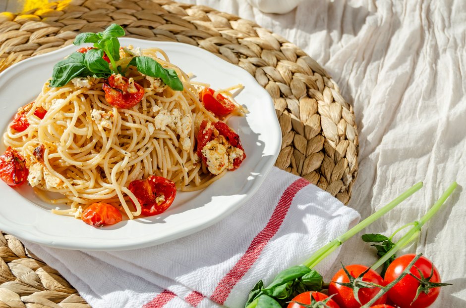 Teller mit Spaghetti, Feta, getrockneten Tomaten und Basilikum auf einem geflochtenen Untersetzer neben frischen Tomaten an Stielen auf einem weißen Tuch mit roten Streifen