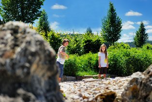 Zwei Kinder spielen draußen auf einem sonnigen Weg mit Bäumen und grünem Buschwerk im Hintergrund
