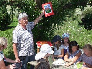 Kinder sitzen im Naturgarten im Weidenpavillion und lernen von einem Lehrer, der ein Buch hält.