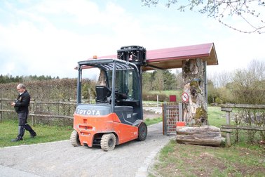Oranger Gabelstapler hebt ein Holzdach über einem Torbogen aus Baumstämmen an, Mann geht daneben auf einem Weg