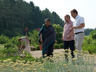 Zwei Männer und eine Frau in einem langen Gewand stehen an einem Blumenbeet und begutachten die farbenfrohen Blumen.