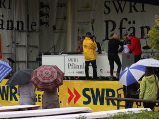 Bühne des SWR Pfännle-Events mit Teilnehmern unter Regenschirmen.