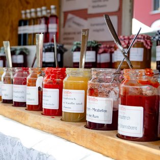 Mehrere Marmeladengläser mit verschiedenen Sorten und Löffeln stehen in einer Reihe auf einem Holzbalken auf einem Marktstand