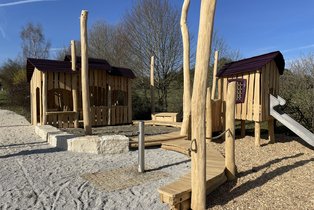 Kinderspielplatz mit zwei Holzhäusern, Rutsche und Kletterelementen auf Kiesboden unter blauem Himmel