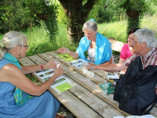 Vier Erwachsene sitzen im Weidenpavillion im Naturgarten bei ALB-GOLD mit verschiedenen Materialien auf dem Tisch.